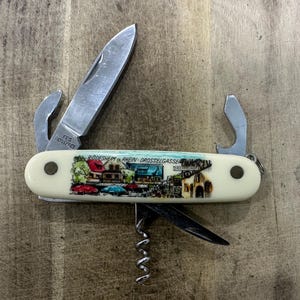 May include: A multi-tool pocket knife with an off-white handle featuring a colourful graphic of a street scene and the text RÜDESHEIM AM RHEIN-DROSSELGASSE. The stainless steel tool includes a main blade marked ROSTFREI, a corkscrew, and bottle opener.