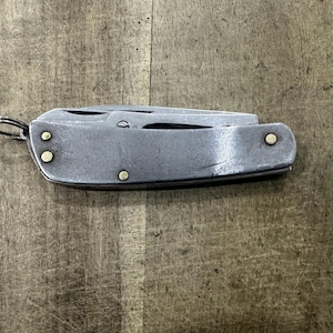 Puede incluir: Una navaja cerrada con mango de metal desgastado que presenta tres pequeños remaches de latón y una anilla de metal en un extremo. La hoja está completamente retraída en el mango, mostrando una textura superficial lisa y usada.