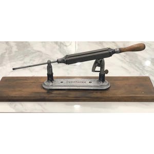 May include: An antique knife sharpener with a wooden handle and a metal frame. The sharpener is mounted on a wooden base and has a long, thin rod extending from the frame. The base has the text "VOORHEES" engraved on it.