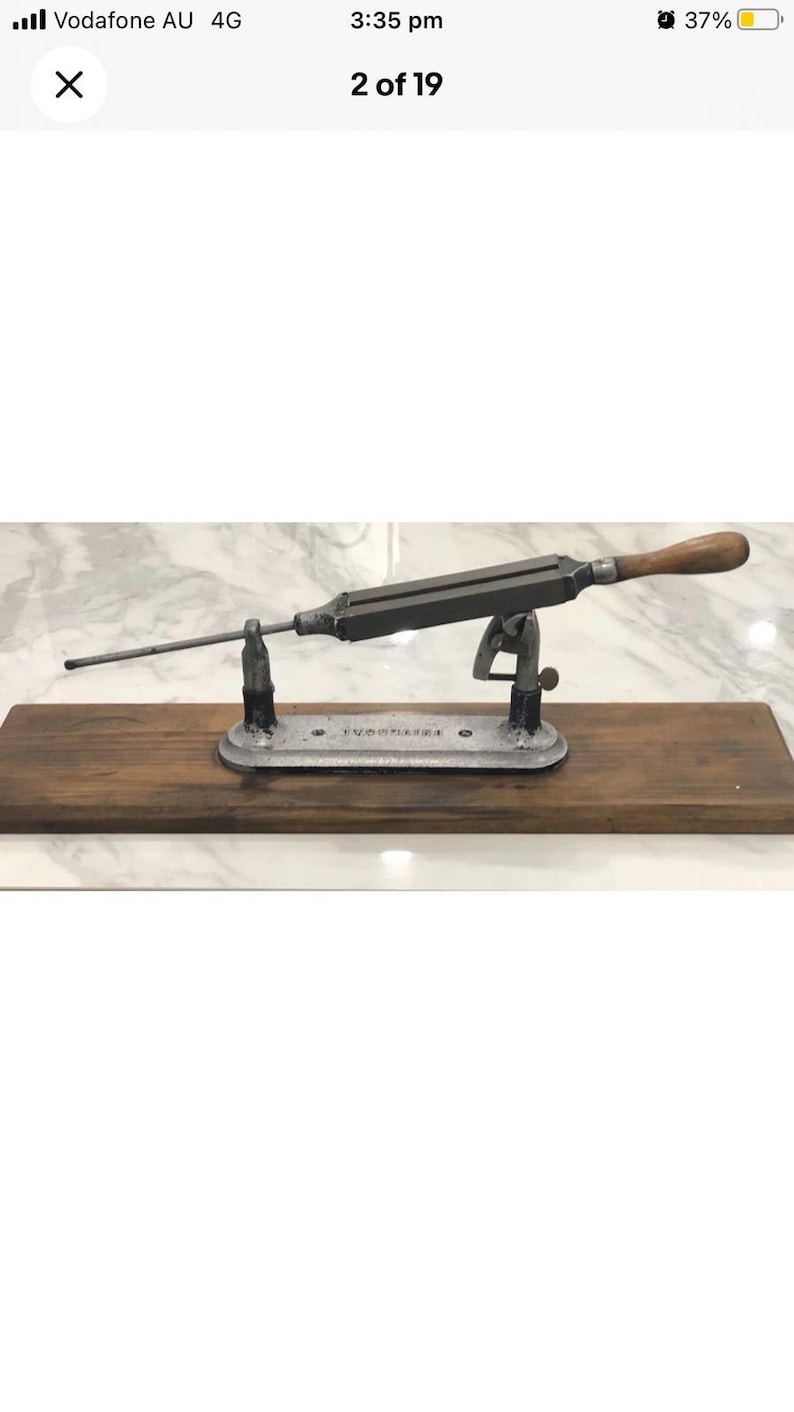 May include: An antique knife sharpener with a wooden handle and a metal frame. The sharpener is mounted on a wooden base and has a long, thin rod extending from the frame. The base has the text "VOORHEES" engraved on it.