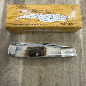 Peut inclure: Un couteau de poche pliant avec un manche texturé aux tons variés et des mitres en métal poli, présenté avec sa boîte en carton jaune d'origine. La boîte porte les inscriptions Parker Eagle Brand Knives, Genuine Sandbar Handles et NO.1423.