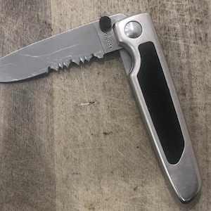Pode incluir: Uma faca dobr&aacute;vel parcialmente aberta com uma l&acirc;mina serrilhada. A faca tem uma al&ccedil;a prateada com uma pega preta texturizada. A l&acirc;mina &eacute; marcada com o texto "Kershaw".