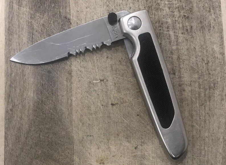 Pode incluir: Uma faca dobr&aacute;vel parcialmente aberta com uma l&acirc;mina serrilhada. A faca tem uma al&ccedil;a prateada com uma pega preta texturizada. A l&acirc;mina &eacute; marcada com o texto "Kershaw".