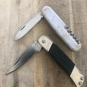 May include: Two pocket knives on a wooden surface. One has a white handle with a corkscrew, the other a black and green handle. The silver blades are partially open, showing the sharp edge of the knives.