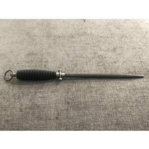 May include: A black and silver knife sharpening steel. The handle is black with a ridged design, and the steel is a slender, tapered rod. A small silver ring is at the end of the handle.