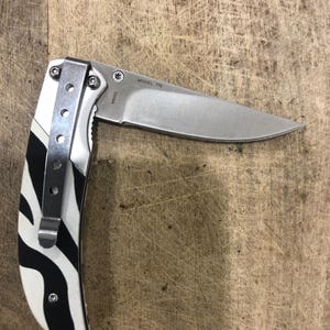 K&ouml;nnte beinhalten: Ein Klappmesser aus Edelstahl mit teilweise ge&ouml;ffneter Klinge. Der Griff hat ein schwarz-wei&szlig;es Zebrastreifenmuster. Das Messer hat einen silbernen Taschenclip und der Text "MODEL 776" ist auf der Klinge sichtbar.