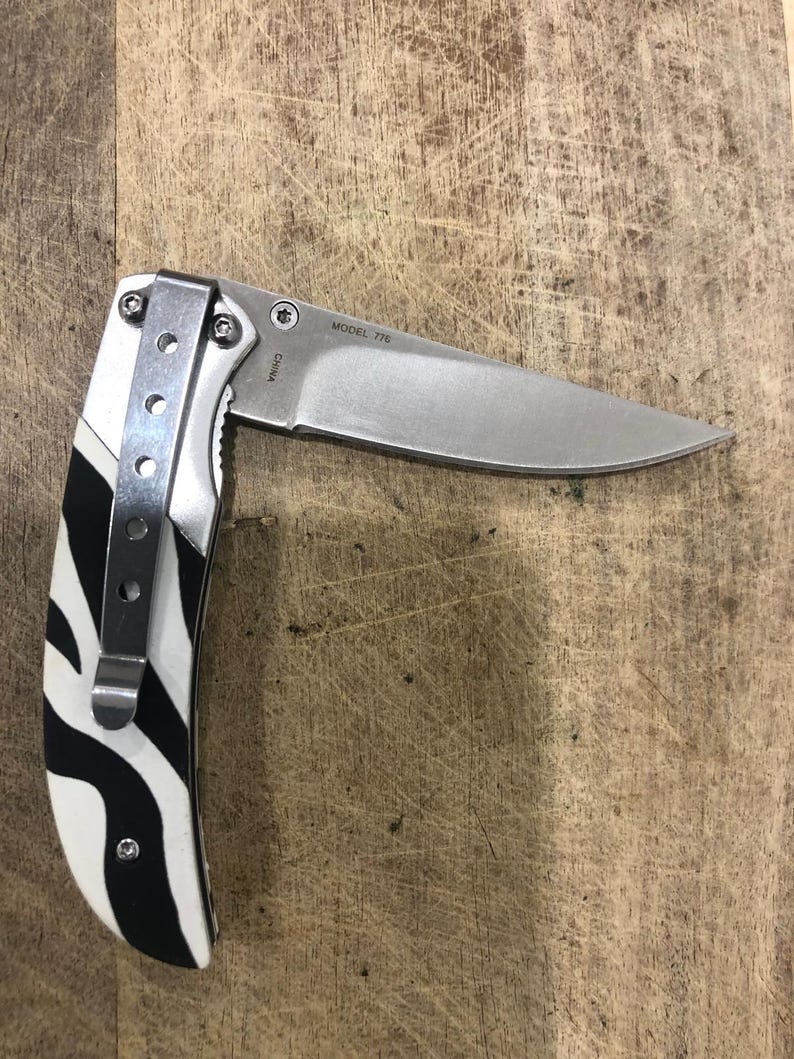 K&ouml;nnte beinhalten: Ein Klappmesser aus Edelstahl mit teilweise ge&ouml;ffneter Klinge. Der Griff hat ein schwarz-wei&szlig;es Zebrastreifenmuster. Das Messer hat einen silbernen Taschenclip und der Text "MODEL 776" ist auf der Klinge sichtbar.