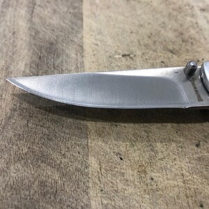 K&ouml;nnte beinhalten: Ein silbernes Klappmesser mit einer scharfen, spitzen Klinge. Der Griff ist silberfarben mit einem rosa Hirschlogo. Die Klinge besteht aus Edelstahl. Das Messer liegt auf einer Holzoberfl&auml;che.