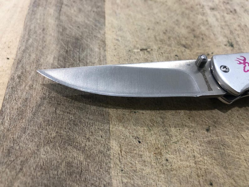K&ouml;nnte beinhalten: Ein silbernes Klappmesser mit einer scharfen, spitzen Klinge. Der Griff ist silberfarben mit einem rosa Hirschlogo. Die Klinge besteht aus Edelstahl. Das Messer liegt auf einer Holzoberfl&auml;che.