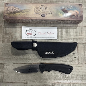 May include: A Buck fixed-blade knife with a black handle and silver blade, a black nylon sheath with a silver snap button, and a rectangular box featuring an elk illustration and text reading 35th Anniversary and RMEF, plus a thank you note.