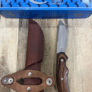 May include: A Colt Ridge Runner skinner knife with a silver stainless steel blade and a dark brown wood grain handle, accompanied by a brown leather sheath with a snap closure and a blue Colt branded box with the text The Legend Continues.