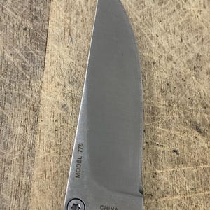 K&ouml;nnte beinhalten: Nahaufnahme einer Edelstahl-Messerklinge mit gebogener Kante. Der Text "MODEL 776" und "CHINA" ist auf der Klinge sichtbar. Das Messer liegt auf einer Holzoberfl&auml;che.