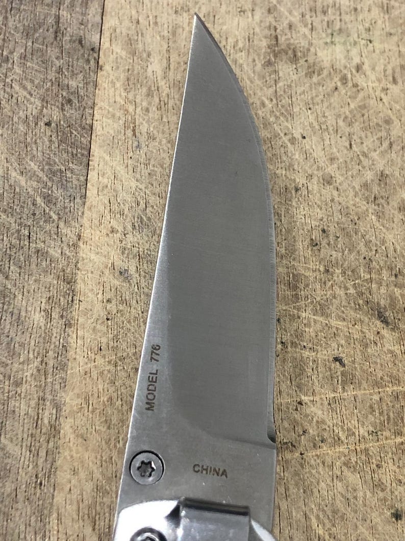 K&ouml;nnte beinhalten: Nahaufnahme einer Edelstahl-Messerklinge mit gebogener Kante. Der Text "MODEL 776" und "CHINA" ist auf der Klinge sichtbar. Das Messer liegt auf einer Holzoberfl&auml;che.