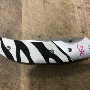 K&ouml;nnte beinhalten: Ein Klappmesser mit einem Zebrastreifenmuster auf dem Griff. Der Griff ist wei&szlig; mit schwarzen Streifen. Die Klinge ist silberfarben und das Messer hat ein rosa Logo auf dem Griff.