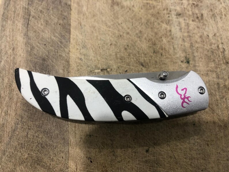 K&ouml;nnte beinhalten: Ein Klappmesser mit einem Zebrastreifenmuster auf dem Griff. Der Griff ist wei&szlig; mit schwarzen Streifen. Die Klinge ist silberfarben und das Messer hat ein rosa Logo auf dem Griff.