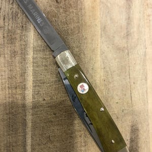 Op de afbeelding: Een vouwmes met een groene handgreep en een zilveren lemmet. Het lemmet is gegraveerd met "U.S. MARINES" en een logo. De handgreep heeft een klein embleem met een rood, wit en blauw ontwerp.