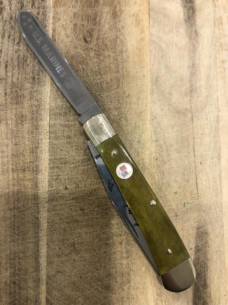 Op de afbeelding: Een vouwmes met een groene handgreep en een zilveren lemmet. Het lemmet is gegraveerd met "U.S. MARINES" en een logo. De handgreep heeft een klein embleem met een rood, wit en blauw ontwerp.