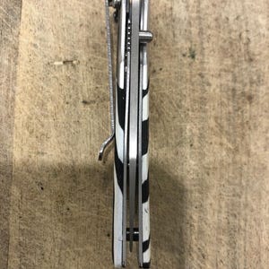 K&ouml;nnte beinhalten: Ein geschlossenes Taschenmesser mit einem schwarz-wei&szlig;en Zebrastreifenmuster auf dem Griff. Das Messer hat einen silbernen Metallclip und eine silberne Klinge. Das Messer ist geschlossen und scheint neu zu sein.