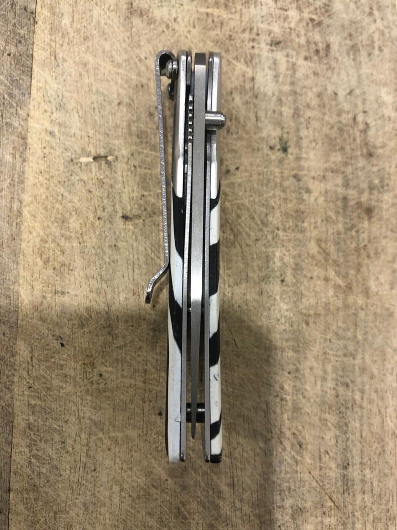 K&ouml;nnte beinhalten: Ein geschlossenes Taschenmesser mit einem schwarz-wei&szlig;en Zebrastreifenmuster auf dem Griff. Das Messer hat einen silbernen Metallclip und eine silberne Klinge. Das Messer ist geschlossen und scheint neu zu sein.