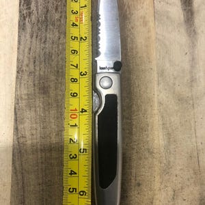 May include: A silver folding knife with a black handle, next to a yellow measuring tape. The knife blade is 7.6 cm long. The measuring tape shows inches and fractions of an inch.