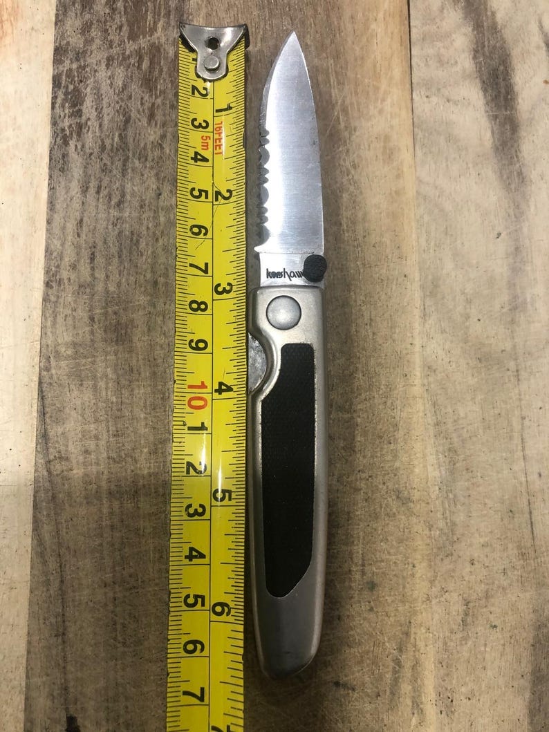 May include: A silver folding knife with a black handle, next to a yellow measuring tape. The knife blade is 7.6 cm long. The measuring tape shows inches and fractions of an inch.