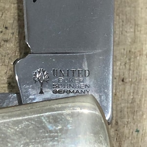 Op de afbeelding: Close-up van een roestvrijstalen vouwmes. Het lemmet is gegraveerd met de woorden "UNITED BOKER SOLINGEN GERMANY" en een boomlogo. De handgreep heeft een glanzende, zilveren kleur.