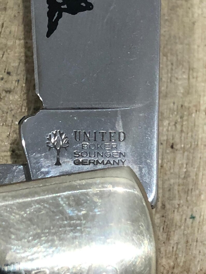 Op de afbeelding: Close-up van een roestvrijstalen vouwmes. Het lemmet is gegraveerd met de woorden "UNITED BOKER SOLINGEN GERMANY" en een boomlogo. De handgreep heeft een glanzende, zilveren kleur.