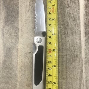 Pode incluir: Uma faca dobr&aacute;vel prateada com uma l&acirc;mina serrilhada, ao lado de uma fita m&eacute;trica amarela. A faca tem uma pega preta e mede aproximadamente 15 cm de comprimento. A marca "Kershaw" &eacute; vis&iacute;vel na l&acirc;mina.