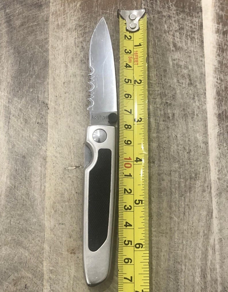 Pode incluir: Uma faca dobr&aacute;vel prateada com uma l&acirc;mina serrilhada, ao lado de uma fita m&eacute;trica amarela. A faca tem uma pega preta e mede aproximadamente 15 cm de comprimento. A marca "Kershaw" &eacute; vis&iacute;vel na l&acirc;mina.