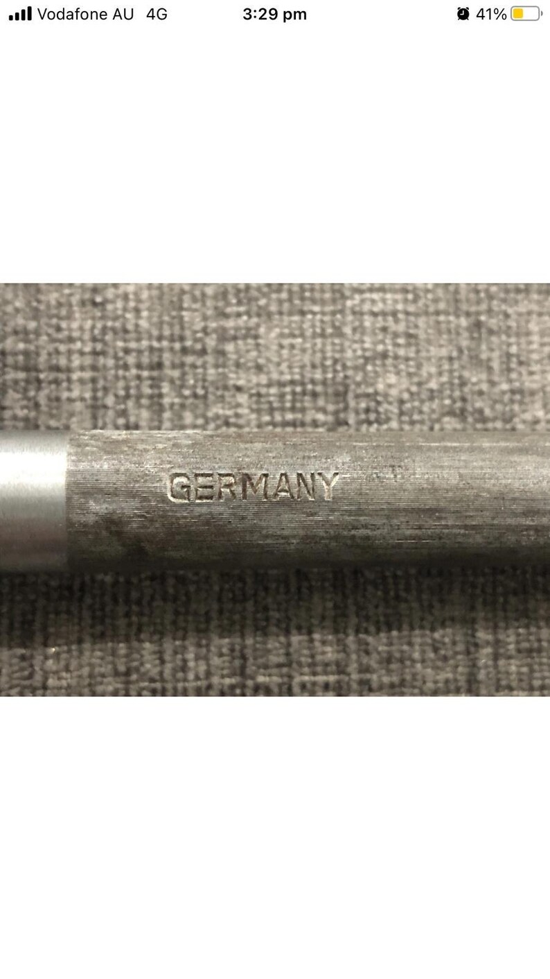 May include: Close-up of a metal object with the word "GERMANY" stamped on it. The object has a silver end and a textured, gray body. The background is a textured, gray surface.