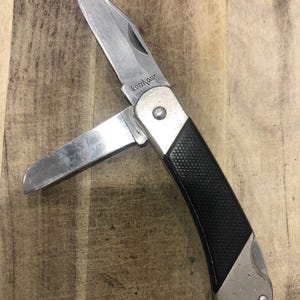 Puede incluir: Una navaja plateada con una hoja parcialmente abierta. El mango es negro con un agarre texturizado y detalles plateados. La marca "Kershaw" es visible en la hoja. La navaja est&aacute; sobre un fondo de madera.