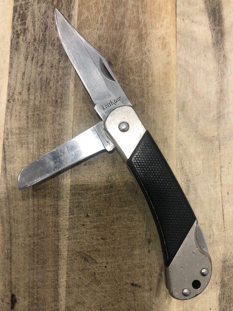 Puede incluir: Una navaja plateada con una hoja parcialmente abierta. El mango es negro con un agarre texturizado y detalles plateados. La marca "Kershaw" es visible en la hoja. La navaja est&aacute; sobre un fondo de madera.