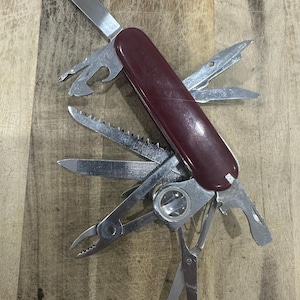 May include: A multi-tool pocket knife with a dark maroon handle and multiple stainless steel implements extended, including a main blade, saw, file, scissors, pliers, a magnifying glass, and various screwdrivers and bottle openers.