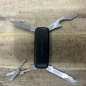 May include: A black multi-tool with a textured handle featuring the embossed text 'SINCE 1893 USA'. The tool has four extended stainless steel implements: a small knife blade, a bottle opener, a pair of scissors, and a flat-head screwdriver tip.