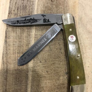 Op de afbeelding: Een vouwmes met een groene handgreep en zilveren accenten. Het lemmet heeft een ge&euml;tste afbeelding van een militaire sc&egrave;ne en de woorden "U.S. MARINES". Een rood-wit-blauw embleem staat op de handgreep.