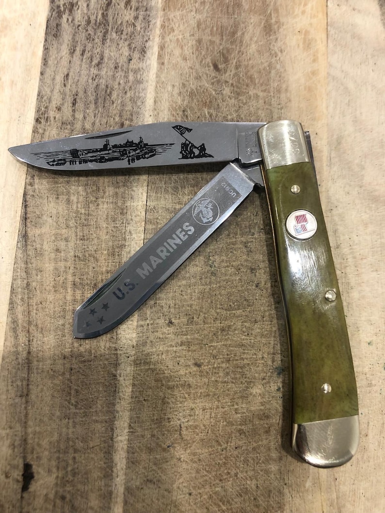 Op de afbeelding: Een vouwmes met een groene handgreep en zilveren accenten. Het lemmet heeft een ge&euml;tste afbeelding van een militaire sc&egrave;ne en de woorden "U.S. MARINES". Een rood-wit-blauw embleem staat op de handgreep.