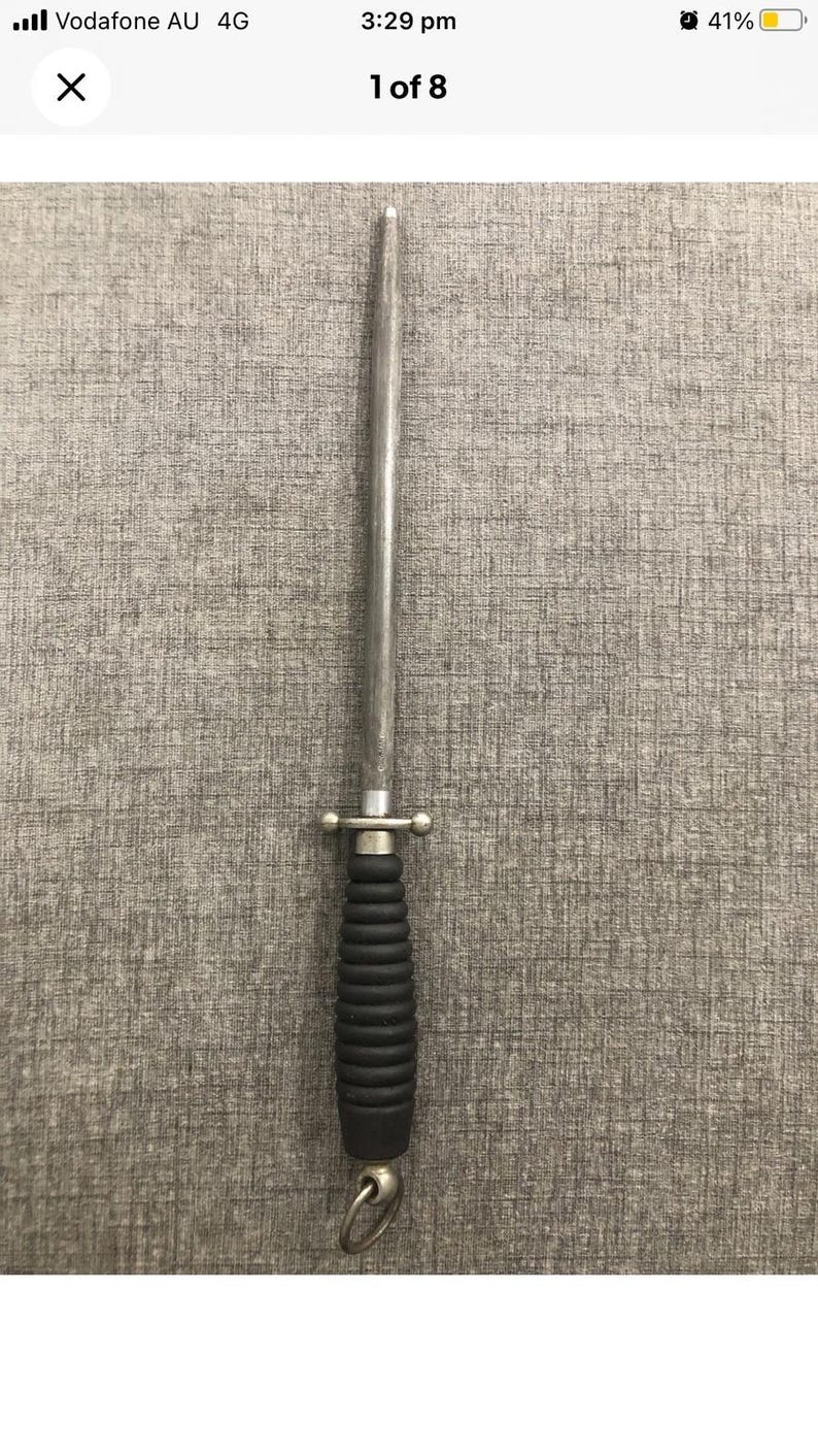 May include: A silver steel knife sharpener with a black, ridged handle and a small metal loop at the base. The sharpener has a long, tapered steel rod and a decorative metal guard.