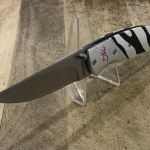 K&ouml;nnte beinhalten: Ein Klappmesser mit silberfarbenem Griff und einem schwarz-wei&szlig;en Zebrastreifenmuster. Die Klinge ist dunkelgrau. Das Messer hat ein rosa Logo und wird auf einem transparenten Kunststoffst&auml;nder pr&auml;sentiert.