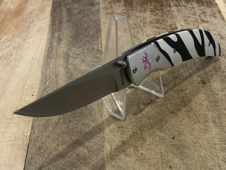 K&ouml;nnte beinhalten: Ein Klappmesser mit silberfarbenem Griff und einem schwarz-wei&szlig;en Zebrastreifenmuster. Die Klinge ist dunkelgrau. Das Messer hat ein rosa Logo und wird auf einem transparenten Kunststoffst&auml;nder pr&auml;sentiert.