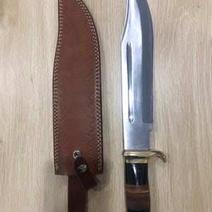 May include: A large hunting knife with a polished steel blade and a handle featuring black, brown, and gold accents. The knife is displayed next to a brown leather sheath with decorative stitching.