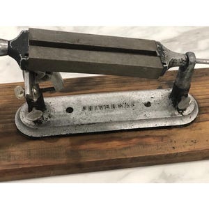 May include: An antique metal Universal knife sharpener mounted on a wooden base. The sharpener has a rectangular sharpening stone and a metal frame. The base is made of dark brown wood. The text "UNIVERSAL" is visible on the metal base.