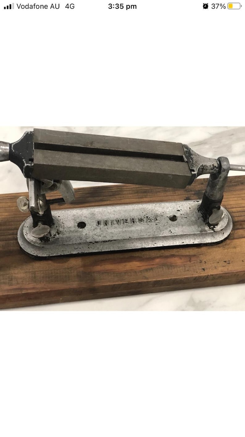 May include: An antique metal Universal knife sharpener mounted on a wooden base. The sharpener has a rectangular sharpening stone and a metal frame. The base is made of dark brown wood. The text "UNIVERSAL" is visible on the metal base.