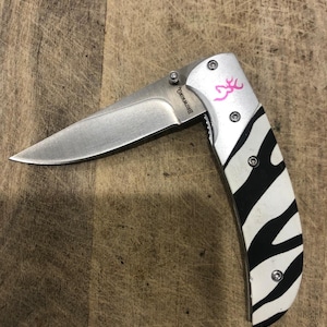 K&ouml;nnte beinhalten: Ein Klappmesser mit einer Edelstahlklinge und einem Griff mit einem Zebra-Streifen-Design in Schwarz und Wei&szlig;. Der Griff hat auch ein rosa Logo und das Wort "Browning" ist auf der Klinge aufgedruckt.