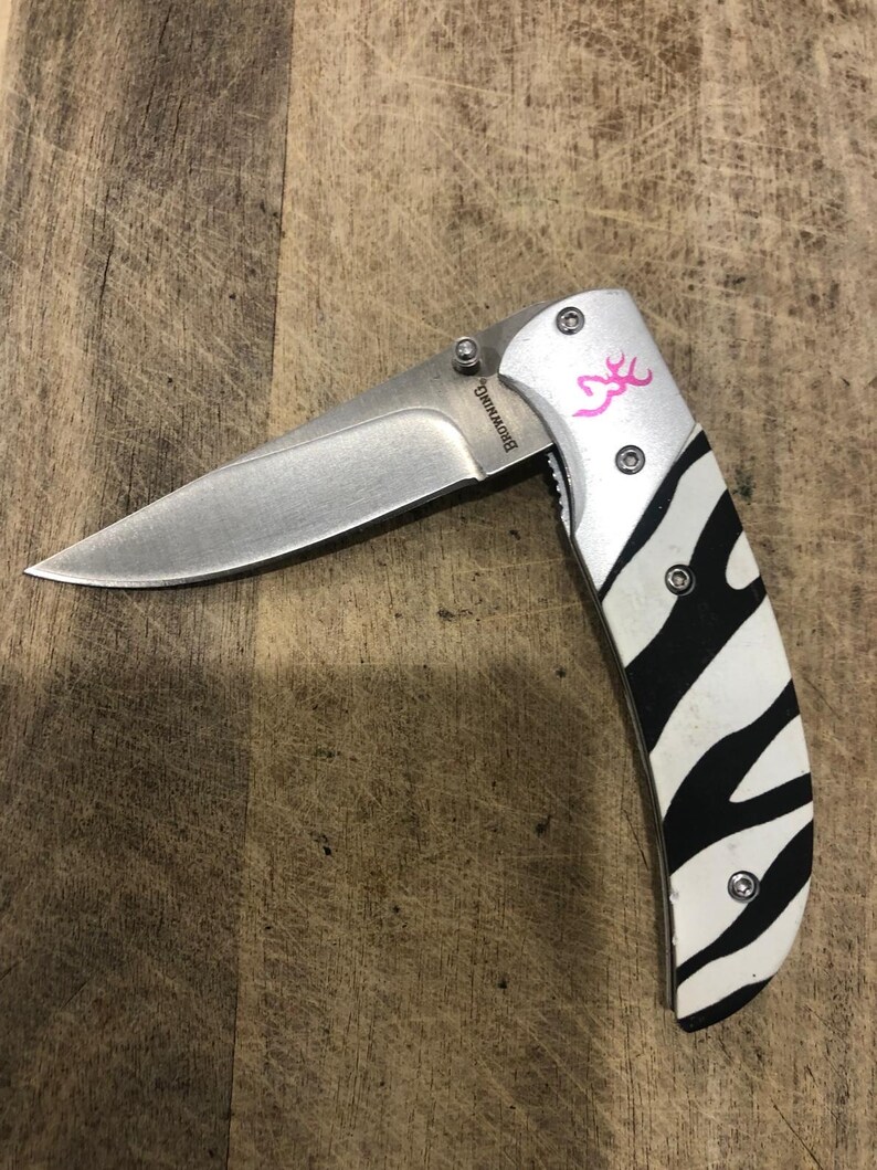 K&ouml;nnte beinhalten: Ein Klappmesser mit einer Edelstahlklinge und einem Griff mit einem Zebra-Streifen-Design in Schwarz und Wei&szlig;. Der Griff hat auch ein rosa Logo und das Wort "Browning" ist auf der Klinge aufgedruckt.