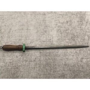 May include: A knife sharpening steel with a dark wooden handle, a green ring, and a steel rod. The sharpening steel is used to maintain the sharpness of knives. The handle has a small loop for hanging.