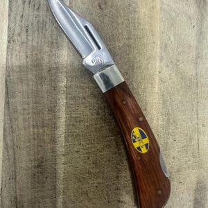 May include: A folding pocket knife with a silver-toned metal blade and a dark brown wooden handle. The blade is stamped with the text CASE XX USA. The handle features a small oval emblem with a yellow and blue flag design and the text HOSSBERG.