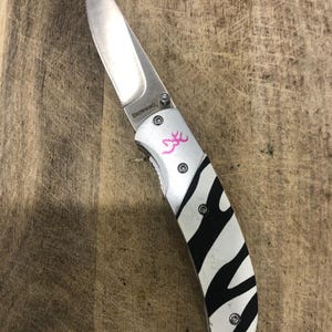 K&ouml;nnte beinhalten: Ein Klappmesser mit einer Edelstahlklinge und einem Griff mit einem schwarz-wei&szlig;en Zebrastreifenmuster. Das Messer hat ein rosa Logo und das Wort "Browning" ist auf der Klinge aufgedruckt.