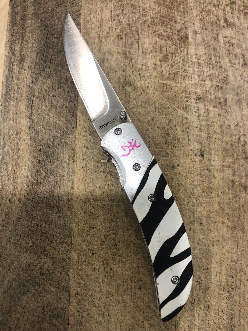 K&ouml;nnte beinhalten: Ein Klappmesser mit einer Edelstahlklinge und einem Griff mit einem schwarz-wei&szlig;en Zebrastreifenmuster. Das Messer hat ein rosa Logo und das Wort "Browning" ist auf der Klinge aufgedruckt.