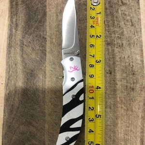 K&ouml;nnte beinhalten: Ein Klappmesser mit einer Klinge aus rostfreiem Stahl und einem Griff mit Zebrastreifen-Design. Der Griff hat ein rosa Logo und die Klinge ist etwa 9 cm lang. Das Messer liegt neben einem Ma&szlig;band.