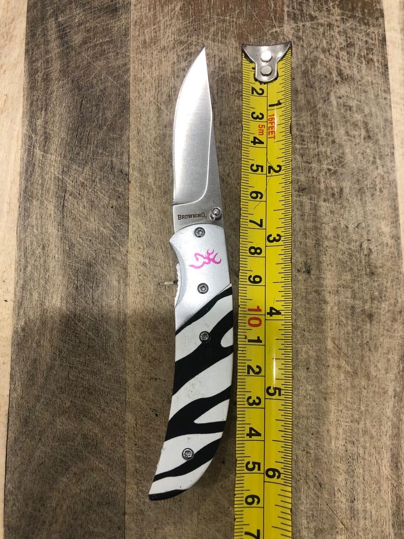 K&ouml;nnte beinhalten: Ein Klappmesser mit einer Klinge aus rostfreiem Stahl und einem Griff mit Zebrastreifen-Design. Der Griff hat ein rosa Logo und die Klinge ist etwa 9 cm lang. Das Messer liegt neben einem Ma&szlig;band.